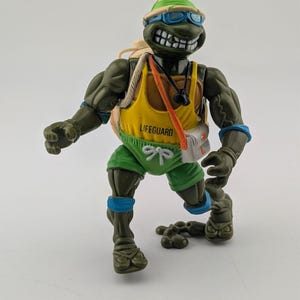 May include: A Teenage Mutant Ninja Turtle action figure in a lifeguard outfit. The figure is green with a yellow tank top that says "Lifeguard", green shorts, and a tan hat. It has blue sunglasses and a white bag.