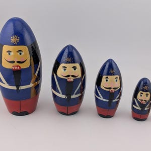 May include: A set of five wooden nesting dolls painted as nutcracker soldiers. Each doll features a blue uniform with white cross-straps, a red base, a black beard, and a pointed blue hat with a gold star emblem on the front of the hat.