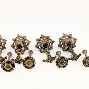 May include: A collection of four ornate brass-toned metal drawer pulls featuring a star-shaped backplate with a drop handle and four matching circular knobs with a raised floral pattern, all finished in a dark, weathered antique bronze tone.