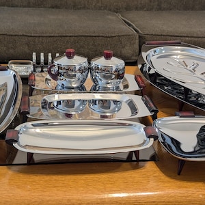 May include: A collection of polished chrome serving pieces featuring dark reddish-brown accents on the handles and lids. The set includes several rectangular and oval trays, two lidded containers, and a large platter with a leaf-like embossed pattern.