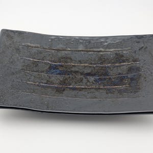 May include: A rectangular glass tray with a dark, glossy black finish. The surface features a textured, uneven centre section with four horizontal, slightly raised lines and subtle hints of deep blue and metallic brown tones within the glass.