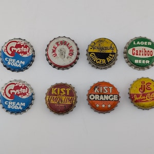 Może przedstawiać: Osiem metalowych kapsli vintage ułożonych w dwa rzędy. Kapsle zawierają różne logotypy marek i napisy, w tym Crystal Cream Soda, Unexcelled, Restigouche Ginger Ale, Cariboo Lager Beer, Kist Lemon Lime, Kist Orange oraz John Collins.