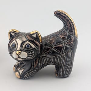 May include: A small ceramic cat figurine featuring a black and metallic bronze finish. The cat has a white muzzle, blue eyes, and gold-toned accents on the ears, nose, and tail. The body is decorated with geometric triangle patterns and textured vertical lines.