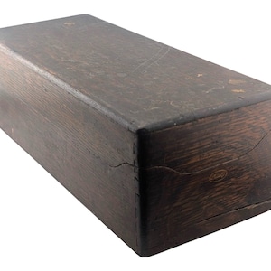 May include: A rectangular dark brown wooden box with a visible wood grain texture. The box features a hinged lid with a curved cut-out design on the front panel and a small, oval-shaped metallic emblem embedded on the lower right side of the front.