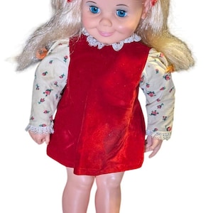 Puede incluir: Muñeca vintage con cabello rubio y ojos azules, vestida con un vestido de terciopelo rojo sobre una blusa con estampado floral. La muñeca tiene zapatos rojos y es un juguete clásico.