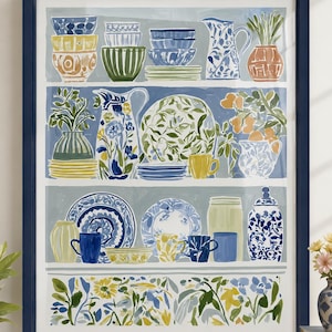 May include: A framed wall art print featuring a three-tiered shelf display of assorted ceramic kitchenware including bowls, pitchers, plates, and mugs in blue, yellow, green, and white, with a floral and botanical border at the bottom.