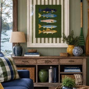 May include: A framed wall art piece featuring five stylized fish in blue, orange, and yellow hues against a dark green background, set within a white frame with a striped mat. The artwork is displayed above a wooden console table with various decor.