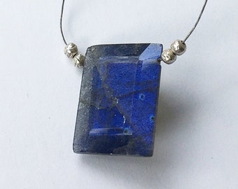 May include: A rectangular, faceted, deep blue gemstone pendant strung on a thin, silver-colored wire. Small, silver-colored beads are placed on either side of the gemstone. The pendant has a slightly irregular shape and a polished surface, reflecting light.
