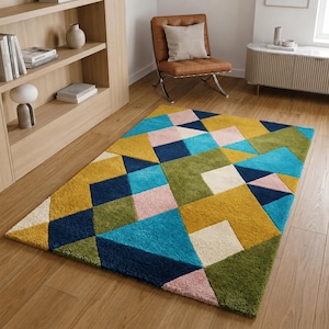 Peut inclure: Un tapis rectangulaire présentant un motif géométrique composé de triangles et de carrés dans des tons jaune moutarde, vert olive, bleu marine, turquoise vif, rose pâle et crème. Le tapis possède une surface en relief touffetée.