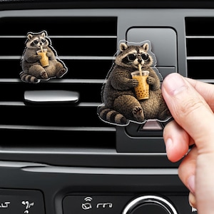 May include: A car air freshener clip featuring a raccoon wearing black sunglasses and drinking a tan bubble tea with a straw. The raccoon has grey and brown fur, a striped tail, and is depicted in a seated position. The design is a flat cutout.