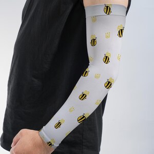 May include: A light gray compression arm sleeve featuring a repeating pattern of small yellow and black bees interspersed with simple yellow line-drawn crowns. The sleeve has a fitted design that covers the forearm and extends to the upper arm.