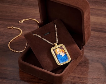 May include: A gold-colored necklace with a rectangular pendant featuring an image of the Virgin Mary and baby Jesus. The pendant is set in a gold frame and rests on a brown velvet display inside a matching box. The necklace chain is also gold.