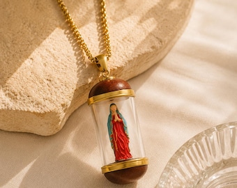 May include: A gold-toned chain necklace featuring a cylindrical pendant. The pendant consists of a clear glass tube capped with dark brown wood at both ends, enclosing a small figurine of a figure in a red robe and blue cloak with hands clasped.
