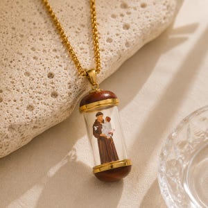 Peut inclure: Un collier à chaîne dorée avec un pendentif cylindrique doté de capuchons en bois foncé et d'un centre en verre transparent. À l'intérieur du cylindre se trouve une figurine détaillée d'un personnage en robe portant un enfant, aux tons brun et blanc.