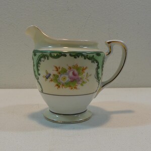 May include: A small white creamer with a green and gold floral design. The creamer has a delicate handle and a rounded base.