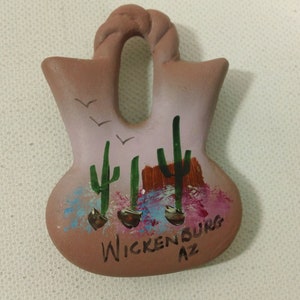 Puede incluir: Un pequeño adorno colgante de cerámica marrón pintado a mano con una escena de tres cactus y pájaros en vuelo. El adorno está etiquetado como "Wickenburg AZ".