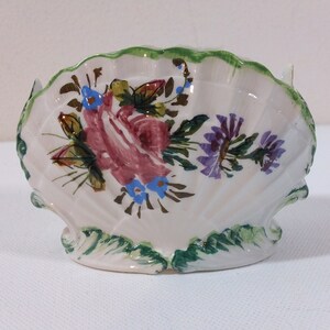 May include: A white ceramic seashell dish with a green rim and painted floral design. The flowers are pink, purple, blue, and green.