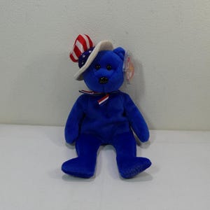 Puede incluir: Un oso de peluche azul con un sombrero blanco y rojo con una cinta blanca y azul alrededor del cuello. El oso tiene una etiqueta roja y blanca pegada a la oreja.