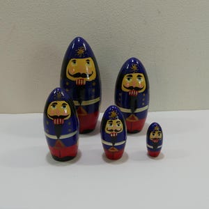 May include: Set of five wooden nesting dolls, each painted with a blue and red nutcracker design. The dolls are arranged in order of size, with the largest doll at the top and the smallest doll at the bottom.