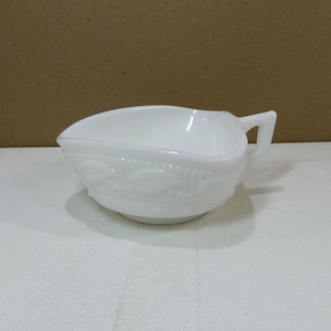 May include: A white, vintage-style creamer or gravy boat. The dish features a spout, a handle, and a decorative embossed pattern around the bowl. The creamer is made of opaque white glass.