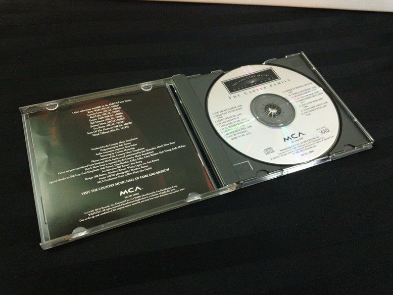 May include: A compact disc case, open to reveal a silver CD with the text "The Country Music Hall of Fame" and "The Carter Family." The case is clear plastic, with a black interior. The CD is labeled with the MCA logo.