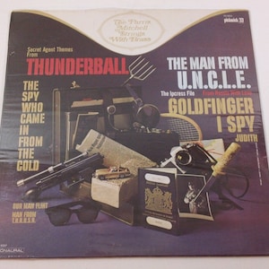May include: A vintage vinyl record cover featuring a collage of spy movie props, including a gun, a passport, sunglasses, and a newspaper. The cover features the titles "Thunderball", "The Spy Who Came in From the Cold", "The Man From U.N.C.L.E.", "The Ipcress File", "Goldfinger", and "I Spy".