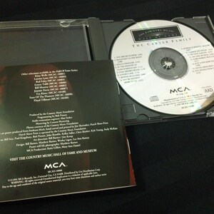 May include: A compact disc (CD) case is open, revealing the CD and its booklet. The CD features the text "The Country Music Hall of Fame" and "The Carter Family." The booklet lists song titles and other information.
