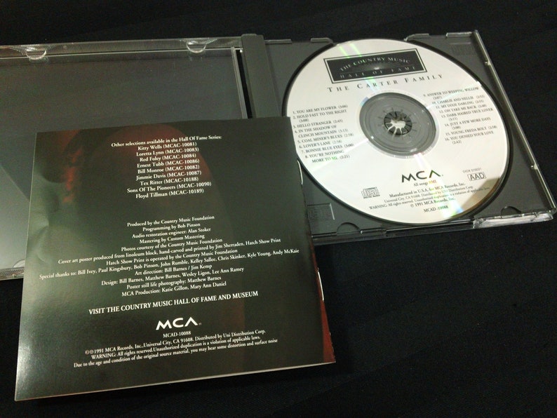 May include: A compact disc (CD) case is open, revealing the CD and its booklet. The CD features the text "The Country Music Hall of Fame" and "The Carter Family." The booklet lists song titles and other information.