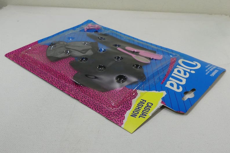 May include: A plastic package with a pink and blue background. The package contains a black and gray outfit for a doll. The package also includes a pink and white comb and a white and pink hairbrush. The package says "Diana Casual Fashion" on the front.