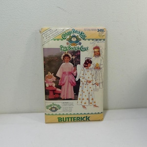 Może przedstawiać: Vintage wzór szycia Cabbage Patch Kids. Wzór zawiera ilustracje dzieci w kostiumach aniołów i wróżek. Opakowanie jest kremowe z logo Butterick na dole. Widoczny jest tekst "Prits Bouts de Choux".