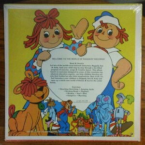 May include: A vintage record album cover featuring Raggedy Ann and Andy, two iconic rag dolls, with a heart-shaped background. The cover is yellow with red polka dots and features a variety of other characters, including a dog, a cat, and a bird. The text on the cover reads "Welcome to the World of Raggedy Records!" and includes a list of exercises for children.