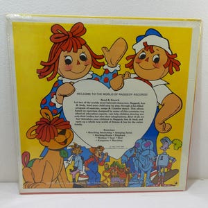 May include: A vintage record album cover featuring Raggedy Ann and Andy, two iconic rag dolls. The cover is yellow with red and blue accents. The text on the cover reads "Welcome to the World of Raggedy Records!" and lists various exercises for children, such as "Reaching, Stretching, Jumping Jacks, Rocking Boats, Elephant, Monkey, Seal, Bird, Kangaroo, Marching."