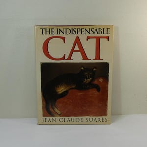 Op de afbeelding: De omslag van het boek "De Onmisbare Kat" toont een zwart-bruine kat die op een rood kussen ligt. De titel "De Onmisbare Kat" is in rode tekst en de auteur "Jean-Claude Suares" is in zwarte tekst.