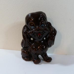 Può includere: Statuetta di cane in ceramica marrone lucido, con occhi blu e bocca rossa. Il cane è seduto, con la testa inclinata e una zampa alzata. La statuetta ha uno stile vintage e da cartone animato.