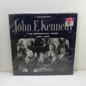 Könnte beinhalten: Ein Schwarzweißfoto von John F. Kennedy, mit dem Text "A Documentary John F. Kennedy The Presidential Years 1960-1963 Recorded by Fox Movietone News" und einem roten Aufkleber mit der Aufschrift "Factory Close Out 57".