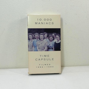 May include: A vintage VHS tape box for the band 10,000 Maniacs, titled "Time Capsule." The box features a black and white photo of several women. The text "Filmed 1982-1990" is printed below the title. The box has a cream-colored background.