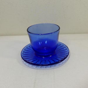 Puede incluir: Un juego de taza y platillo de vidrio azul cobalto. La taza tiene una forma redondeada y se asienta sobre un platillo a juego con un diseño acanalado. El juego se coloca sobre una superficie transparente y texturizada.