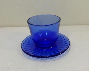 May include: A cobalt blue glass teacup and saucer set. The cup has a rounded shape and sits on a matching saucer with a ridged design. The set is placed on a clear, textured surface.