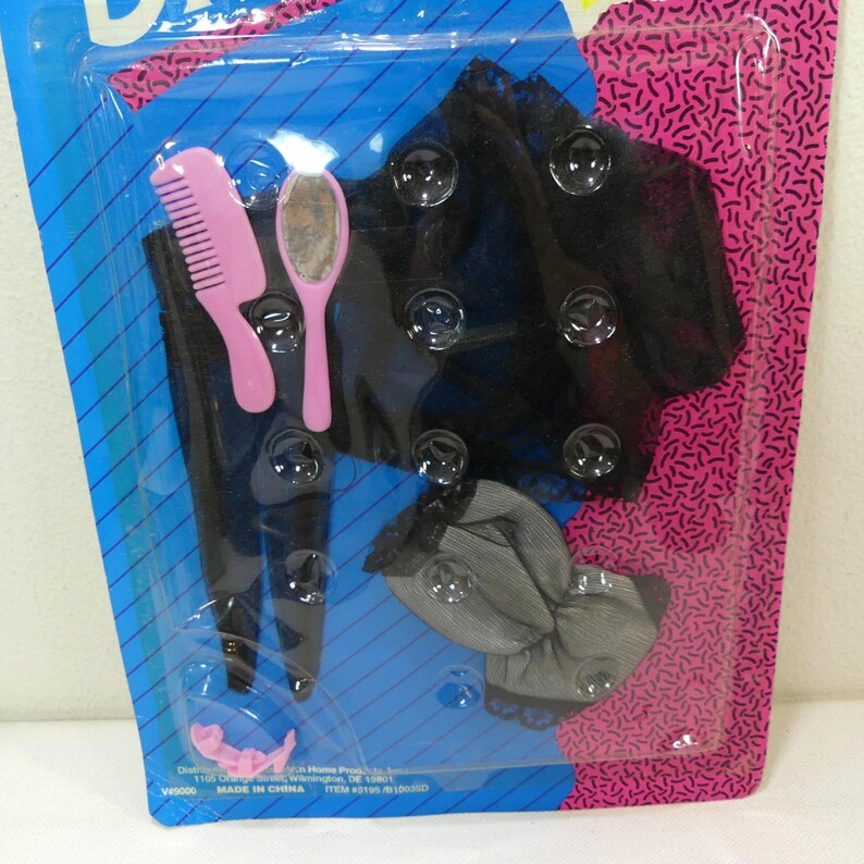 May include: A plastic package containing a black lace dress, a black mesh top, a pink comb, and a pink hand mirror. The package has a blue background with white stripes and a pink background with white squiggles.