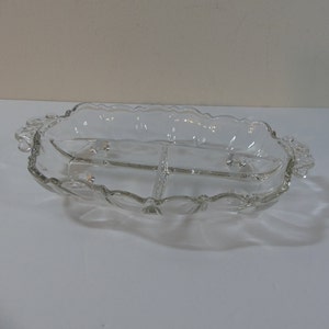 May include: A clear glass serving dish with three divided sections and scalloped edges. The dish has two handles on the sides and a raised base.