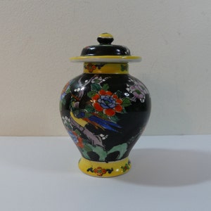 Puede incluir: Un jarrón de cerámica negro con tapa, decorado con diseños florales y de pájaros coloridos. El jarrón tiene un borde y una base amarillos.