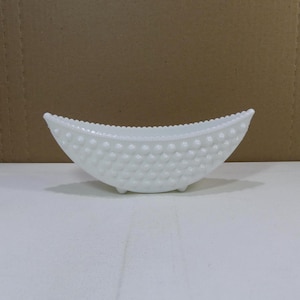 May include: A white, oval-shaped hobnail glass bowl with three small feet. The bowl has a textured surface of small, rounded bumps and a scalloped rim. The bowl is empty and sits on a white surface.