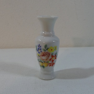 Puede incluir: Un pequeño jarrón de porcelana blanca con un diseño floral. El jarrón tiene un cuello estrecho y una base ancha. El diseño floral presenta una rosa rosa, flores amarillas y naranjas y hojas verdes.