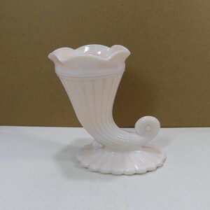 May include: A pink glass vase shaped like a horn with a scalloped rim and a curled handle.