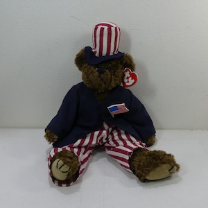 Könnte beinhalten: Ein brauner Teddybär, der einen rot-weiß-gestreiften Hut, eine marineblaue Jacke und rot-weiß-gestreifte Hosen trägt. Der Bär hat eine kleine amerikanische Flaggennadel an seiner Jacke. Der Bär sitzt auf einer weißen Oberfläche.