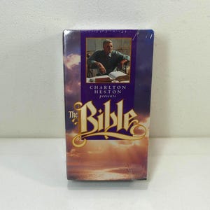 Op de afbeelding: Een VHS-band van "The Bible" met Charlton Heston. De hoes heeft een paarse banner met de titel in gouden letters en een foto van Heston. De achtergrond toont een bewolkte lucht met een zonsondergang.