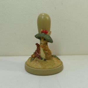 Puede incluir: Figura decorativa con un conejo que lleva un sombrero verde con una flor roja. El conejo sostiene un objeto con estampado rojo y blanco. La figura tiene una base beige y un mango marrón claro.