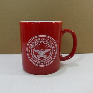 Könnte beinhalten: Rote Keramiktasse mit einem weißen Logo, auf dem "Director of Civilian Marksmanship" mit einem Stern- und Adlerdesign steht.