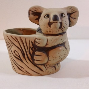 May include: A ceramic koala planter with a brown and tan glaze. The koala is holding onto a tree trunk that forms the planter. The planter is perfect for small plants or succulents.