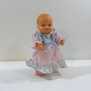 May include: A small plastic doll with blonde hair wearing a pink and white floral dress with a blue bow.